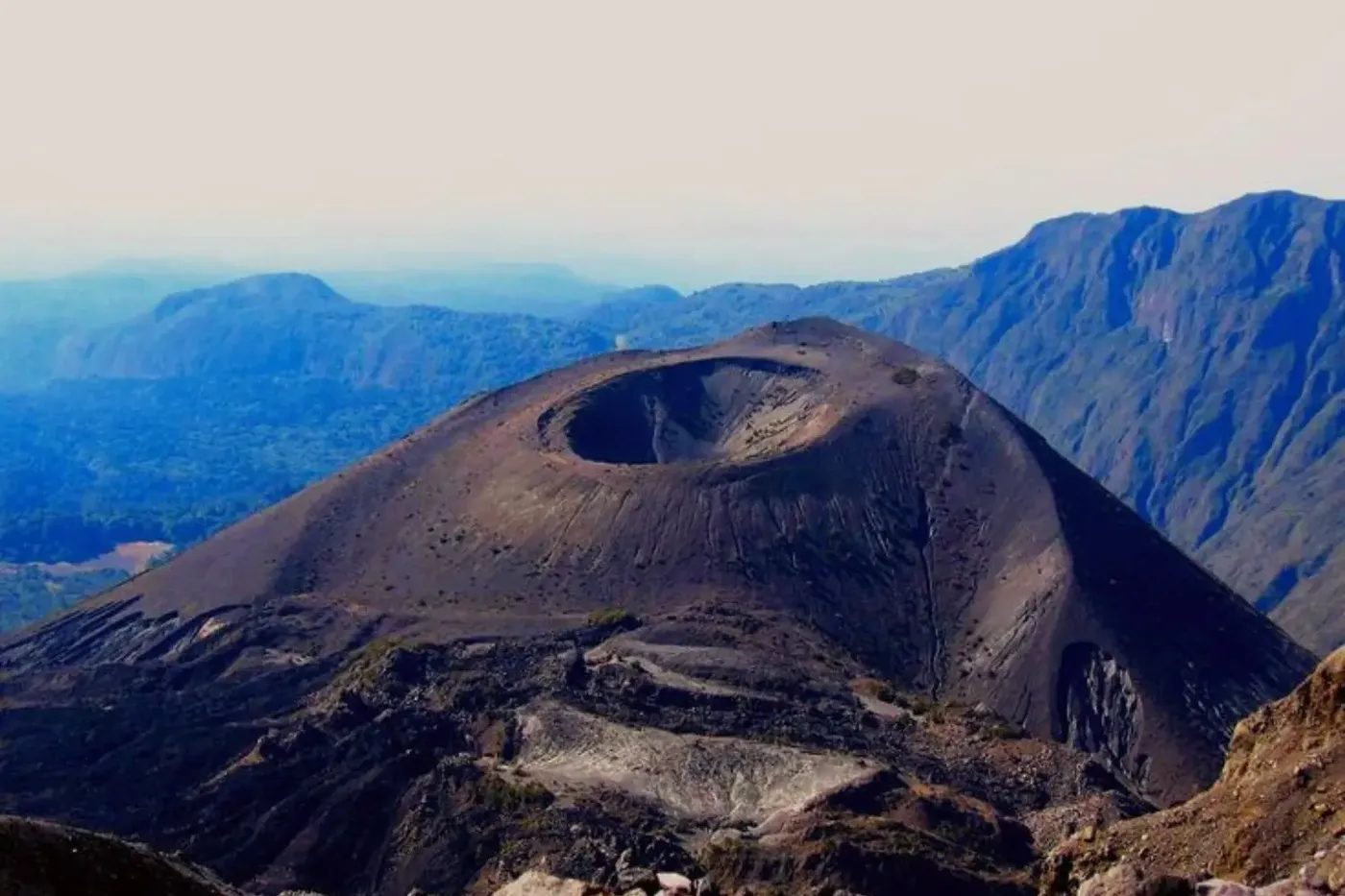 A view of Mount Meru showing its steep volcanic cone and large summit crater, surrounded by rugged dark rock and distant blue mountain ranges beneath a hazy sky.