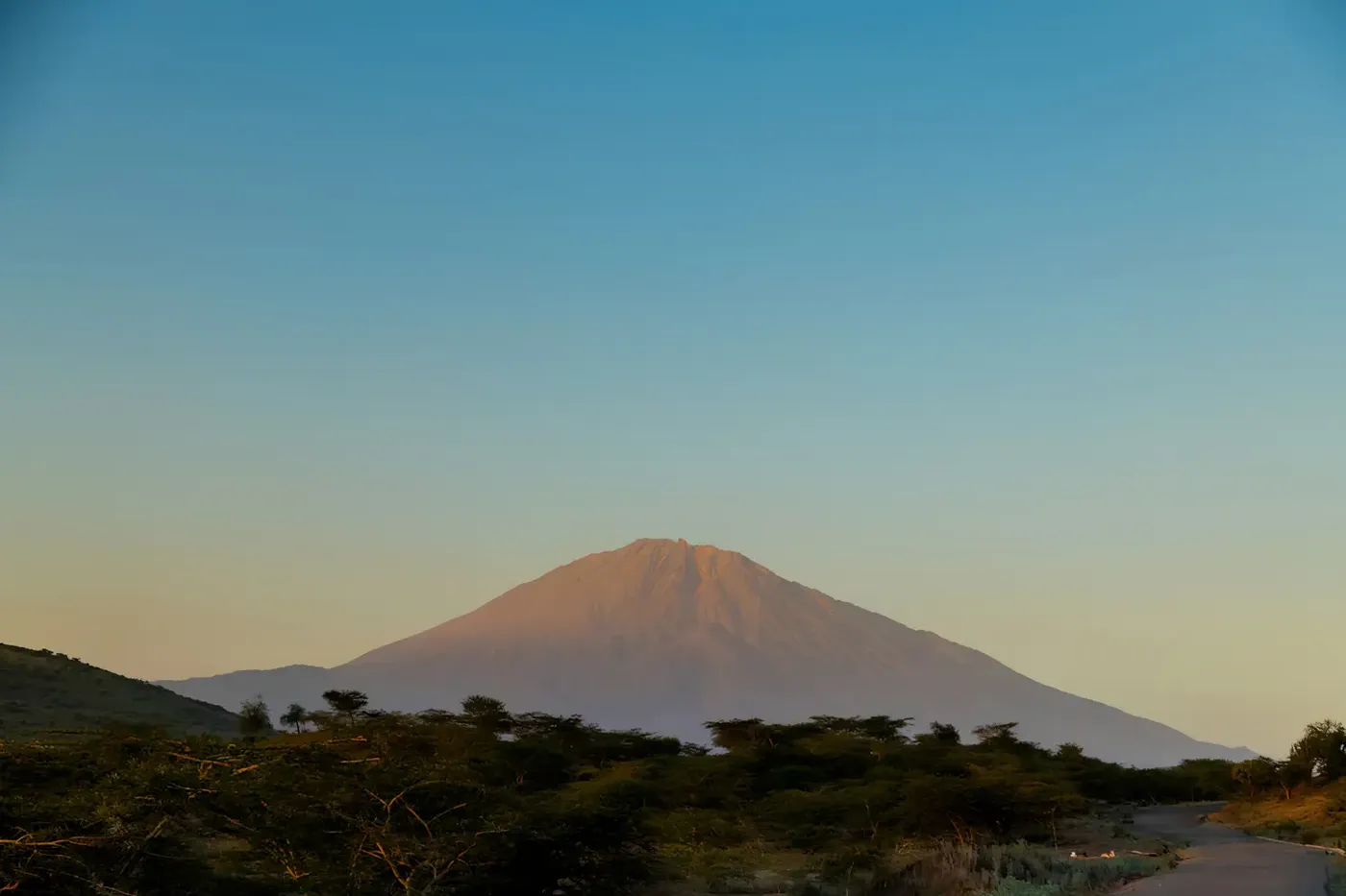 Distant view of Mount Meru rising above the savannah landscape under a clear blue sky at sunrise.
